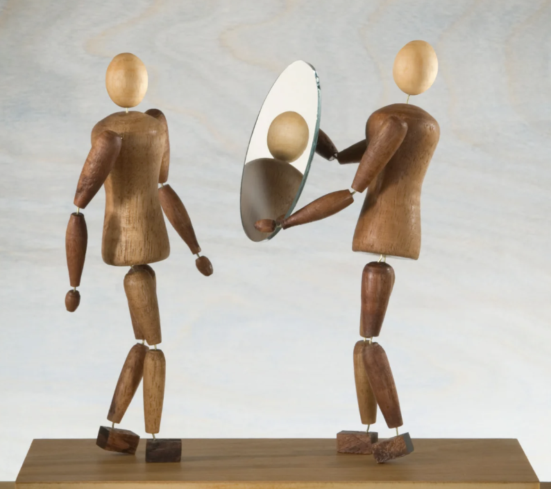 Two wooden figures, one holding a round mirror so the other can see its own reflection